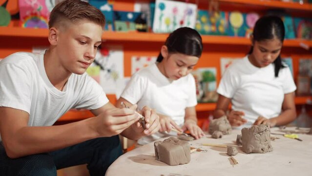 high school teenager using carving tool working at clay at pottery workshop. Group of happy diverse children working or modeling cup of clay with dough and equipment scatter around. Edification