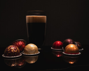 Large black coffee in a double wall glass cup with foam illuminated from above, colourful Nespresso virtue coffee pods reflecting on marble, black background. Still life and product photography.