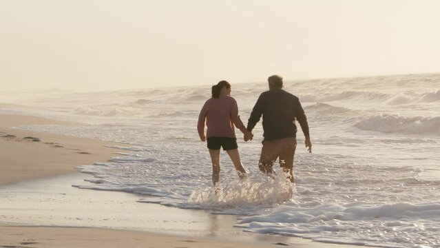 Rear view of loving retired senior couple on vacation running along beach shoreline splashing through waves and holding hands at sunrise - shot in slow motion