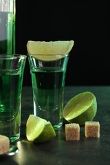 Absinthe in shot glasses, lime and brown sugar cubes on gray table against black background....