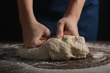 Making bread. Woman kneading dough at wooden table on dark background, closeup