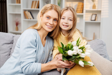 Beautiful mother and daughter holding a bouquet. They are sitting together on sofa in living room