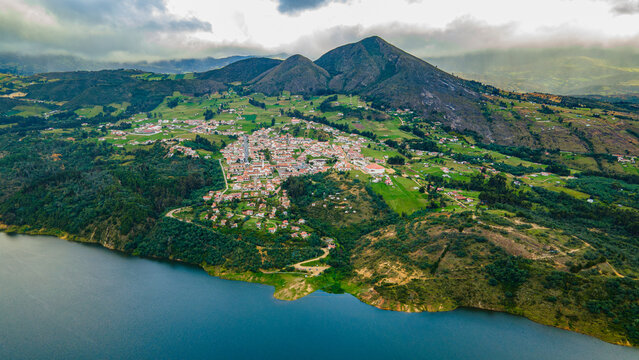 Aerial view of Guatavita town nestled by the tranquil Tomine Reservoir, surrounded by lush green hills under a cloudy sky