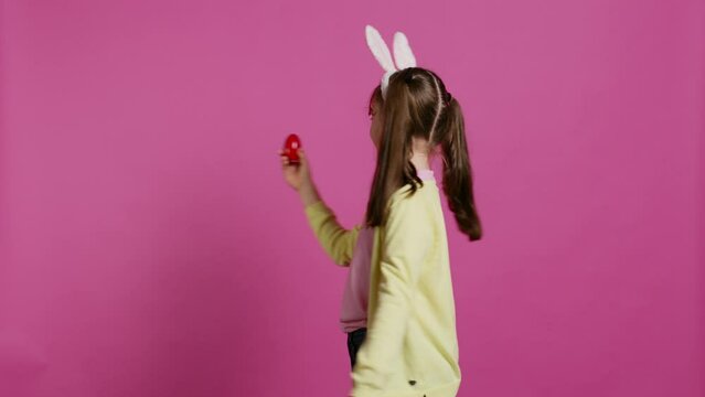 Enthusiastic small child spinning around in front of camera, presenting her red painted egg for easter sunday festivity. Cheery toddler with bunny ears twirling and doing a pirouette. Camera B.