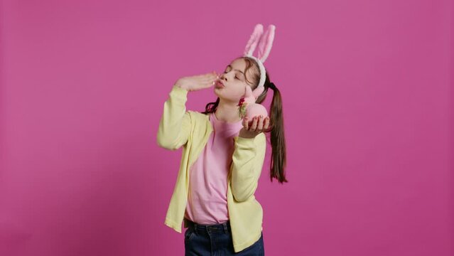Jolly schoolgirl holding a cute rabbit toy and blowing kisses in studio, being adorable with festive decorations for easter holiday event. Happy child wearing bunny ears and pigtails. Camera B.