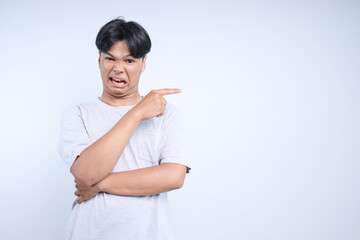 Asian young man pointing aside to the empty space for ads or text and looking at camera with disgusting expression