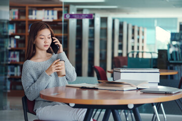 Unhappy young asian woman talking on mobile phone while reading book in library.