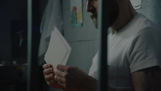 Depressed male prisoner looks at pictures of family or kids sitting on bed in prison cell. Illegally convicted man serves imprisonment term in jail. Detention center or correctional facility. Portrait