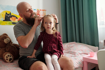 Young bearded man sitting on bed of his adorable blond daughter in smart dress and white tights and braiding her hair
