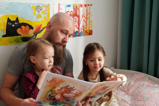 Young bearded man discussing pictures in books of comics with his two adorable daughters while sitting on bed between them