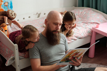 Bearded father holding open book of comics while sitting on the floor by bed with his two adorable...