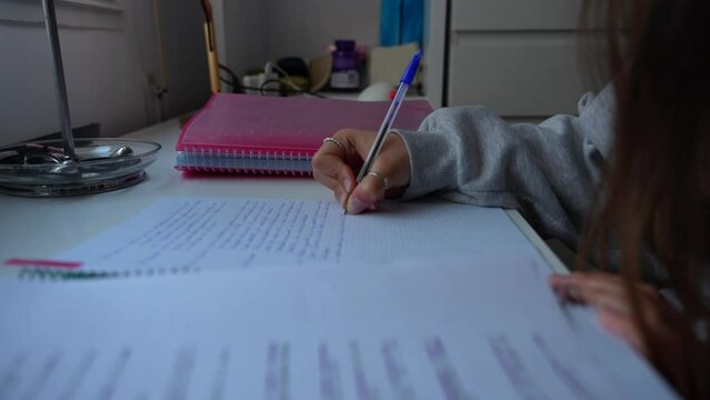 Unrecognizable young girl student taking notes with blue pen on desk by window