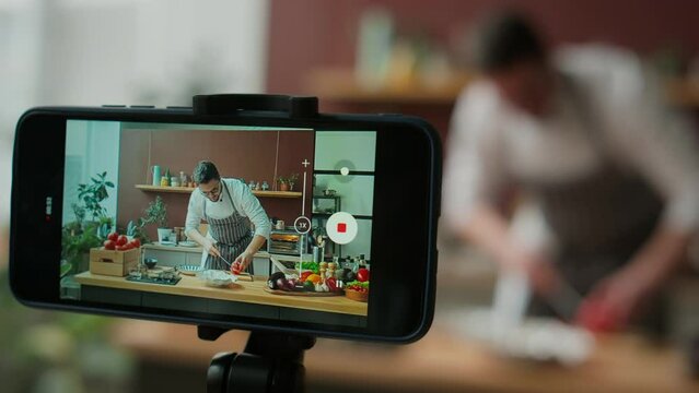 Through smartphone display shot of Caucasian young male chef in workwear filming cooking video in kitchen, demonstrating how to cut grapefruit with knife on wooden board