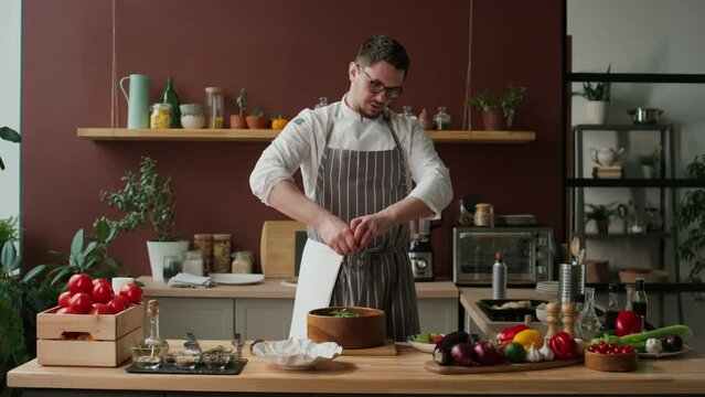 Medium shot of Caucasian young male chef in workwear and eyeglasses squeezing grapefruit juice into salad while recording cooking show in domestic kitchen