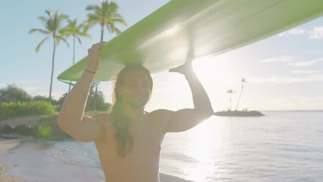 Young barechested male surfer overwhelmed with positive emotions holding surfboard on head, walking along sunny coastline washed by gentle waves of ocean. High quality 4k footage