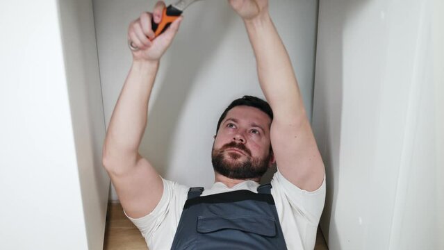 Strong focused repairman lying on floor in t-shirt under overall and tightening valve with wrench while squinting with one eye. Attentive male checking plumbing fixtures for suitability for using.