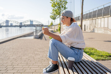 Fototapeta premium Young Hispanic hipster girl browsing mobile phone while sitting in street with takeaway beverage
