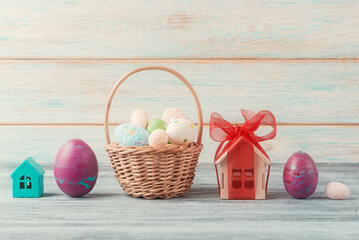 A festive Easter setting featuring a basket of decorated eggs, a miniature turquoise house, and a house-shaped gift box with a red bow - 752747517