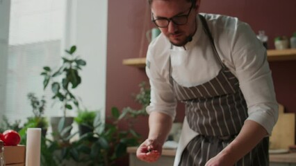 Medium shot of focused young Caucasian male chef with microphone near his mouth adding grapefruit slices to arugula salad and sprinkling seeds on it while cooking meal in bright kitchen