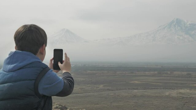 Looking behind him, a teenage boy sitting on the top of a mountain takes pictures of beautiful nature and mountains with majestic and high snowy peaks using a smartphone camera.