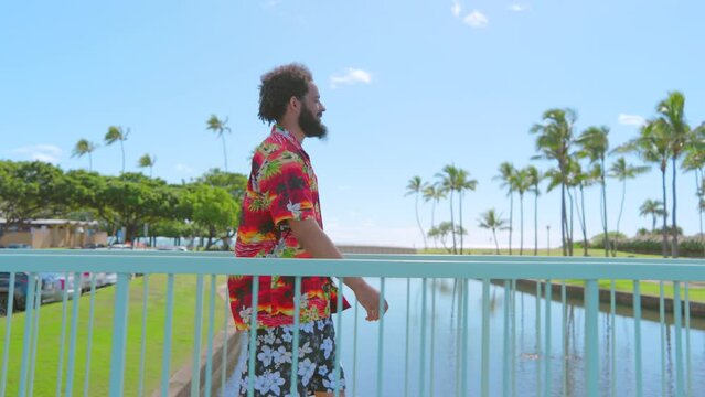 Side view of relaxed dark-haired bearded male tourist in Hawaiian shirt and shorts walking leisurely in a tropical park, crossing bridge over water body. High quality 4k footage