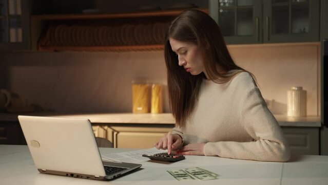 A young woman sets at her kitchen table in the evening, her brows furrowed in concentration as she works on a budget. Surrounded by papers and a laptop, she uses a calculator to manage your finances