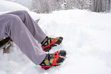 Male legs of a man sitting on a snow covered bench.  The man is wearing gray trousers and mountain boots with Grödel on them.  Grödel, a type of spikes, are used for winter hikes on icy surfaces. - obrazy, fototapety, plakaty