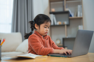 Concentrated Asian young girl studying on laptop at home. Young Asian girl focused on using a laptop for her studies in a comfortable home setting. Education for kid student concept.