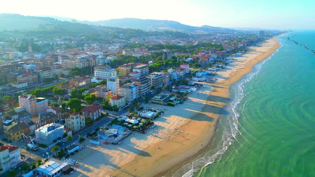 Majestic aerial push-in shot from Porto San Giorgio (Fermo) of the rippling creamy concentrical Adriatic Sea waves washing the sandy shore, with beach establishments, roads, cars, buildings, hillscape