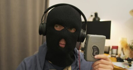 Funny man in a robber mask listens to a retro cassette player with headphones and shakes his head to the beat of the music. Portrait, indoors