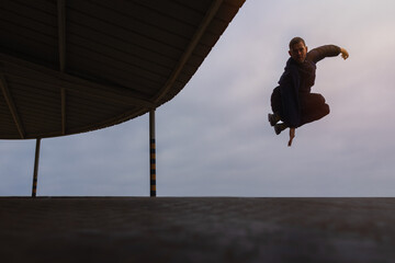 Young man in pants and hoodie jumping on factory ramp. Mid air parkour pose in with overcast sky