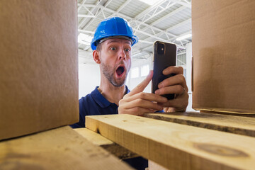 Warehouse worker in helmet shocked expression face when he see result on phone. Stock of parcels with products, transportation concept