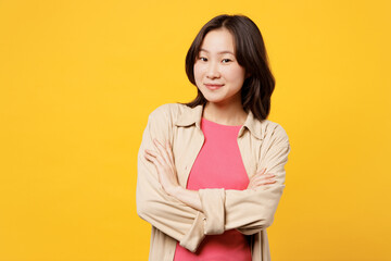 Young smiling happy woman of Asian ethnicity wear pink t-shirt beige shirt pastel casual clothes hold hands crossed folded look camera isolated on plain yellow background studio. Lifestyle portrait.