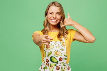 Fototapeta premium Young happy housewife housekeeper chef cook baker woman wear apron yellow t-shirt doing phone gesture like says call me back wink isolated on plain pastel green background studio Cooking food concept