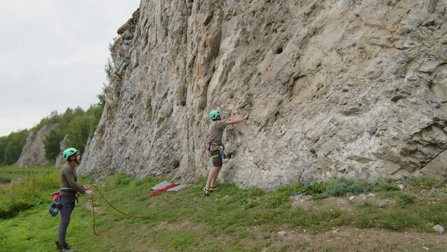 Zoom shot of man climbing up, inserting bolt into rock and attaching rope to quickdraw while his girlfriend standing on ground