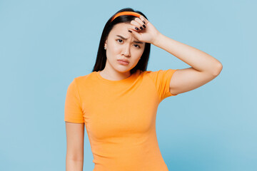 Young fun ill sick sad tired exhausted woman of Asian ethnicity wear orange t-shirt casual clothes put hand on forehead isolated on plain pastel light blue cyan background studio. Lifestyle concept.