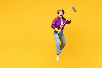 Full body excited young fun woman wear purple shirt casual clothes do housework tidy up jump high hold brush broom jump high isolated on plain yellow background studio portrait. Housekeeping concept.