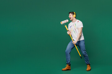 Full body young laborer man wear stained t-shirt headphones hold paint roller pov microphone sing song isolated on plain green background. Instruments renovation apartment room. Repair home concept.
