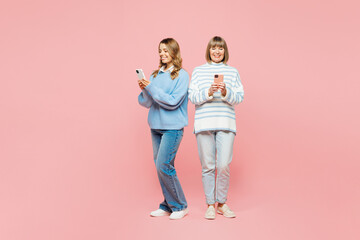 Full body smiling elder parent mom with young adult daughter two women together wear blue casual clothes hold use mobile cell phone isolated on plain pastel light pink background. Family day concept.