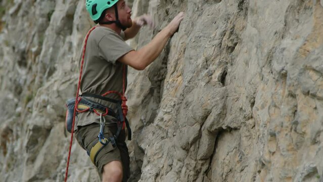 Young male climber wearing protective helmet ascending steep rock and clipping rope to quickdraw