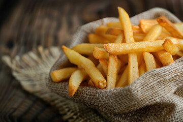 A rustic serving of golden French fries presented in a burlap sack, placed on a vintage wooden tabletop.