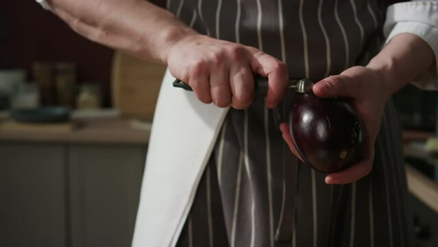 Cropped shot of unrecognizable male chef in striped apron removing pulp from eggplant with stainless steel spoon while cooking in kitchen