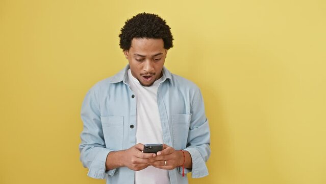 Amazed young african american man, mouth wide open in disbelief, fear and shock, holding his cellphone, standing isolated against a yellow background, expressing wow emotion.