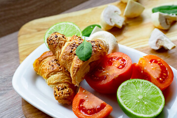 two fresh baked halfs of sliced and stuffed croissant, mushrooms, mint, lime and ripe red halfs of tomato on white ceramic plate. Bakery concept	