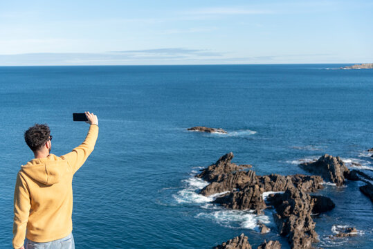 Man with his back turned in a yellow sweatshirt taking a picture of the sea from the cliffs on a sunny day on the Basque coast.