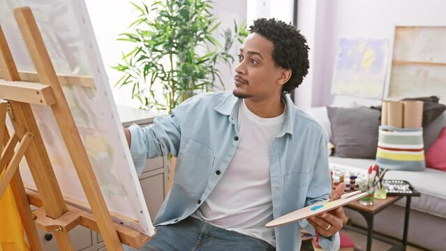 A young, creative, african american man paints on canvas in a well-lit indoor art studio, exuding concentration and talent.