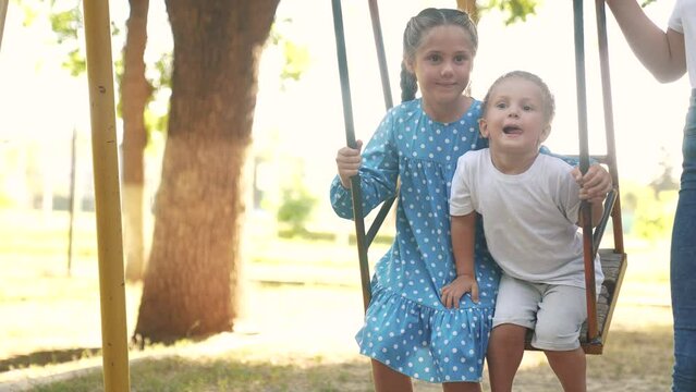 two children on a swing. concept of a happy childhood and loving family. girl and boy in beautiful clothes swings on a swing on the playground, they are lifestyle being filmed by their parents