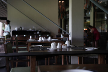 A fine dining or a nice restaurant table view in the morning in a tropical country. A hotel empty breakfast place in the morning with many wooden tables and chairs with white plates and cups.