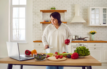 Portrait of fat overweight young woman preparing fresh vegetable salad looking at recipes on a...