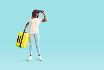 Full length portrait of happy smiling african american woman tourist in casual clothes with suitcase looking through binoculars on studio blue background. Summer holidays trip and vacation concept.